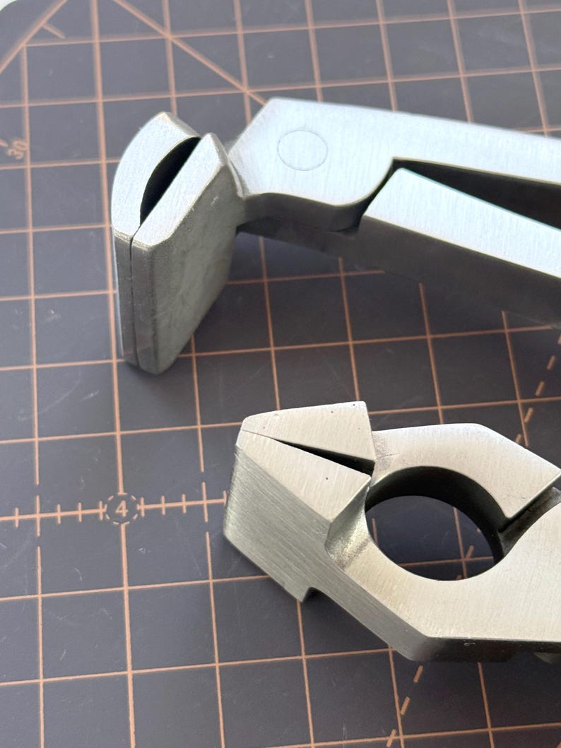 Puede incluir: Primer plano de dos herramientas de metal plateado con un acabado mate. Las herramientas tienen un dise&ntilde;o &uacute;nico con componentes curvos y angulados, lo que sugiere una funci&oacute;n especializada. El fondo es un patr&oacute;n de cuadr&iacute;cula.