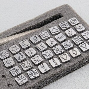 May include: A set of metal stamps with various nature-themed designs, including a butterfly, leaf, and dragonfly. The stamps are arranged in a gray foam insert, with a metal handle visible. The stamps are silver in color.