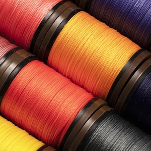 May include: Close-up of colorful spools of thread. The threads are in shades of red, orange, yellow, purple, and black. Each spool has a dark brown and black band around the middle. The threads are tightly wound and create a textured surface.