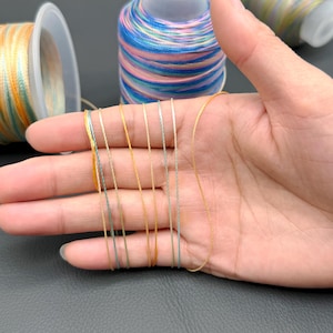 May include: A hand displays several strands of colorful thread in shades of gold, green, and blue. Spools of thread in various colors are visible in the background, suggesting a craft or sewing project. The threads are thin and appear to be made of a shiny material.