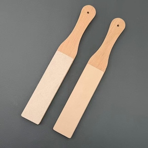 May include: Two wooden stropping paddles with light-coloured leather surfaces attached to the flat blades. Each paddle features a long, smooth wooden handle with a small circular hole at the top end for hanging, showcasing a natural wood grain.