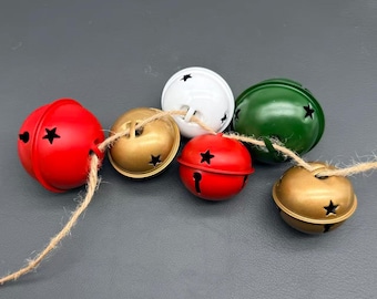 Puede incluir: Un conjunto de seis cascabeles de metal en colores rojo, blanco, verde y dorado, cada uno con aberturas en forma de estrella. Los cascabeles están ensartados en un solo trozo de cuerda de fibra natural marrón, creando una guirnalda decorativa de esferas.