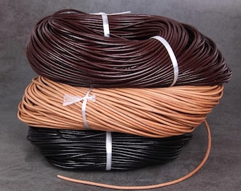 May include: Three spools of leather cord in varying colors: dark brown, tan, and black. Each spool is bound with white ribbon. A single strand of tan cord lies in front of the spools.