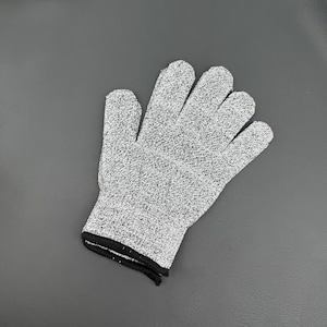 Puede incluir: Un guante resistente a cortes con una construcción de tejido de punto gris y blanco moteado. El guante tiene un diseño flexible con cinco dedos y un puño elástico negro en la muñeca para asegurar un ajuste firme en tareas manuales.