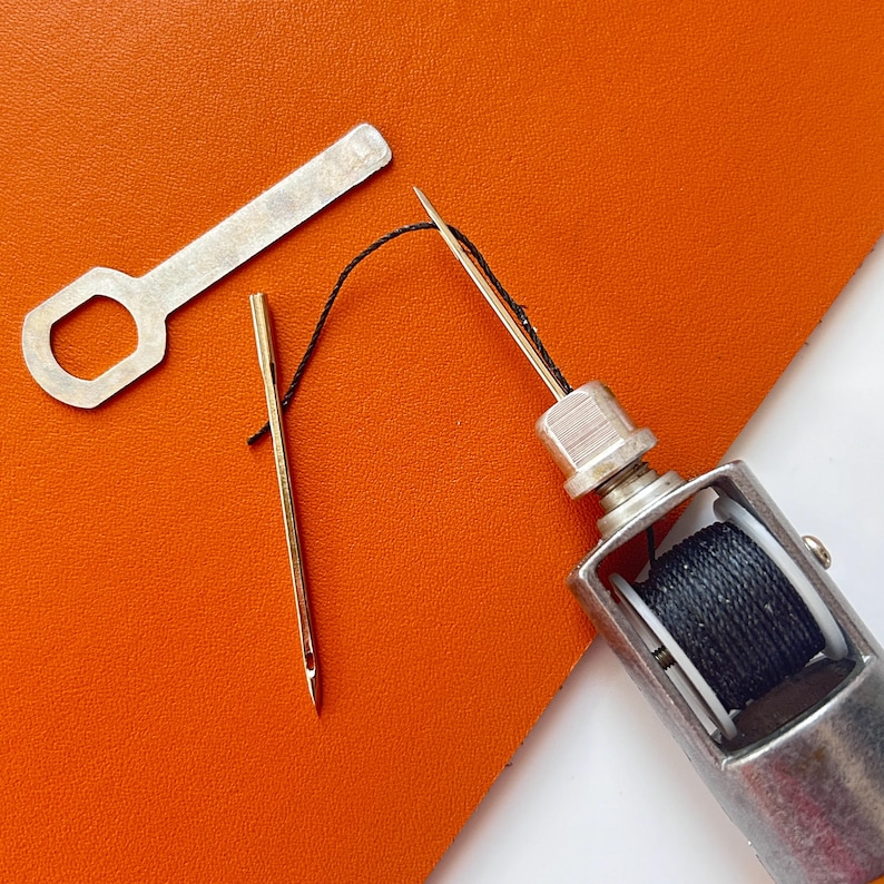 May include: A collection of sewing tools, including a needle threader, a needle, and a thread spool. The tools are silver and gold, with black thread. The background is orange and white.