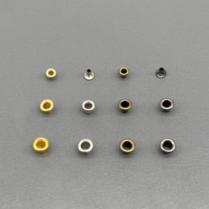 Könnte beinhalten: Eine Sammlung von Metallösen in verschiedenen Farben, darunter Gold, Silber und Bronze. Die Ösen sind auf einer grauen Oberfläche angeordnet und zeigen ihre verschiedenen Ausführungen und Stile. Diese kleinen, kreisförmigen Befestigungselemente werden häufig beim Basteln und in der Bekleidungsherstellung verwendet.