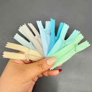 Puede incluir: Una colección de cremalleras coloridas en tonos beige, gris, azul y verde, abanicadas. Cada cremallera tiene un tirador de metal y se muestra sobre un fondo gris. Ideales para proyectos de costura y manualidades.