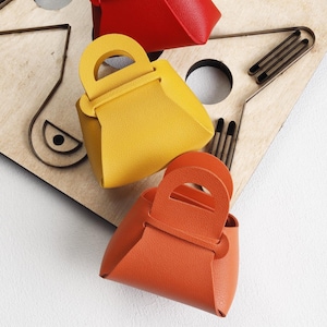 Op de afbeelding: Drie kleine, kleurrijke leren tassen met gebogen handvatten. De tassen zijn rood, geel en oranje en zijn gerangschikt op een lichtgekleurd houten oppervlak met uitgesneden vormen en metalen accenten. De tassen hebben een eenvoudig, geometrisch ontwerp.