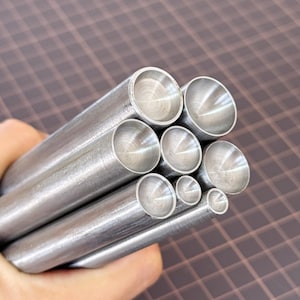 May include: A collection of various sized, silver-colored metal tubes. The tubes are held in a hand and are arranged in a bundle. The ends of the tubes are visible, showing a circular opening. The background is a brown grid.