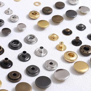 以下が含まれることがあります： シルバー、ゴールド、ブロンズ、ブラックなど、さまざまな色とスタイルの金属製スナップファスナーの詰め合わせ。ファスナーはさまざまなサイズとデザインがあり、衣類やクラフトプロジェクトに適しています。