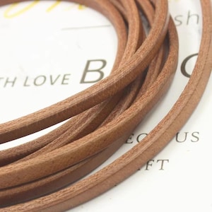 May include: Close-up of several strands of brown leather cord. The cords are tightly coiled and appear to be new. The background is a white surface with the words "WITH LOVE" and the letter "B" visible.