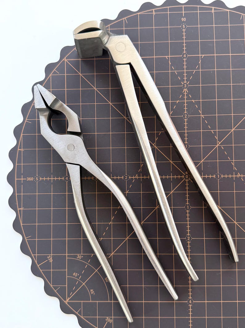 Puede incluir: Dos herramientas de herrero de metal, una tenaza y un martillo de herrar, se muestran sobre una esterilla de corte negra con un patr&oacute;n de cuadr&iacute;cula. La tenaza tiene un dise&ntilde;o similar a unas tijeras con mordazas anguladas. El martillo de herrar tiene una cabeza de martillo en un extremo y una garra en el otro.