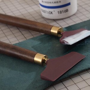 K&ouml;nnte beinhalten: Zwei Lederhandwerkzeuge mit dunkelbraunen Holzgriffen und weinroten Klingen. Die Griffe haben goldfarbene Metallakzente. Ein Werkzeug hat wei&szlig;es Material auf der Klinge. Im Hintergrund befindet sich eine wei&szlig;e Flasche mit blauem Text.