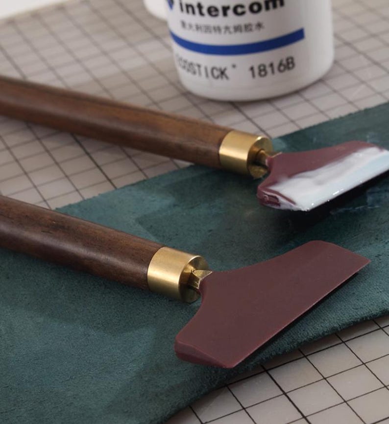 K&ouml;nnte beinhalten: Zwei Lederhandwerkzeuge mit dunkelbraunen Holzgriffen und weinroten Klingen. Die Griffe haben goldfarbene Metallakzente. Ein Werkzeug hat wei&szlig;es Material auf der Klinge. Im Hintergrund befindet sich eine wei&szlig;e Flasche mit blauem Text.