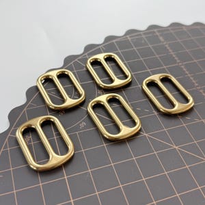May include: Five gold-coloured rectangular belt buckles with a double-bar design. The buckles are made of a shiny metal and are arranged on a dark grey cutting mat with angle markings.
