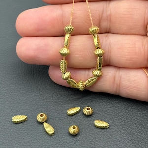 Könnte beinhalten: Goldfarbene Perlenkette mit blatt- und rundförmigen Perlen. Zusätzliche lose Perlen sind darunter verstreut. Die Perlen sind auf einer dünnen, hellfarbigen Schnur aufgefädelt. Die Perlen haben eine strukturierte, metallische Oberfläche.