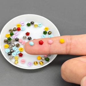 May include: A collection of small, colorful flower-shaped beads in a white dish. The beads are in various colors, including red, yellow, blue, pink, and green. Some beads are on a finger. These beads are likely used for jewelry making or other crafts.