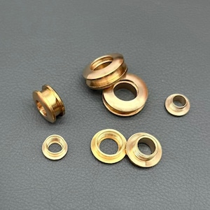 May include: A collection of six metallic brass-colored eyelets and grommets of varying sizes. The components feature circular shapes with central openings, some with threaded inner walls and others with smooth, flanged edges, arranged in a loose cluster.