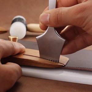 Puede incluir: Primer plano de herramientas de artesanía en cuero en uso. Se utiliza una herramienta metálica con borde dentado sobre cuero marrón oscuro. También se ven otras herramientas, como una herramienta de madera y un mazo.