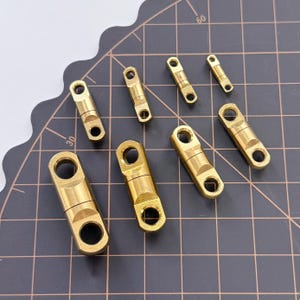 Può includere: Una collezione di girelle da pesca in ottone di varie dimensioni. Le girelle hanno una forma a barilotto con occhielli per attaccare lenze e esche. Sono disposte su un tappetino da taglio nero con linee a griglia.