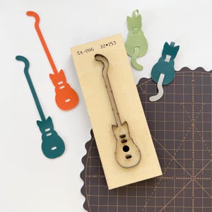 May include: A wooden die-cut guitar shape with the text "SX-086 32*153". Surrounding the die-cut are guitar-shaped leather cutouts in teal, orange, and green. Also, there are cat-shaped leather cutouts in green and teal.
