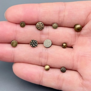 May include: A collection of antique brass-colored metal buttons in various shapes and sizes. The buttons include round, floral, and textured designs. The buttons are displayed on a light background.