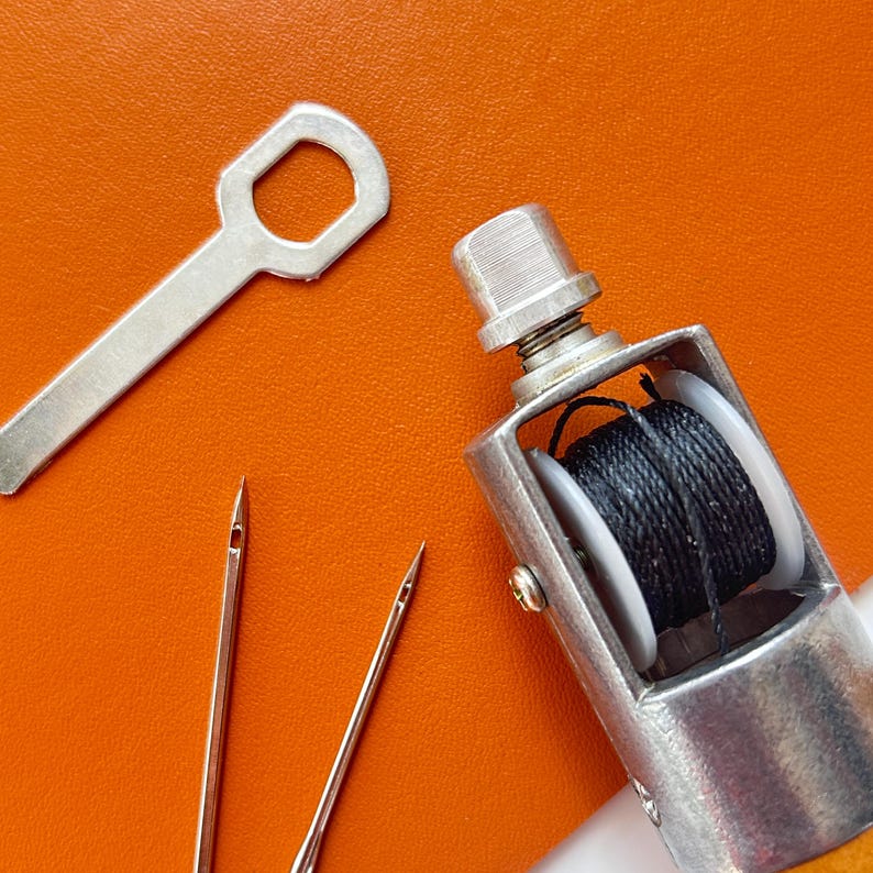 May include: A collection of leatherworking tools, including a silver-colored stitching awl with black thread, two needles, and a silver wrench-like tool. The tools are arranged on an orange surface.