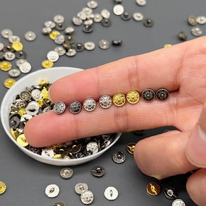 Könnte beinhalten: Eine Auswahl an Metallknöpfen in Silber, Gold und Schwarz. Die Knöpfe haben dekorative Blumenmuster und werden in einer kleinen weißen Schüssel und in einer Hand gezeigt. Die Knöpfe sind auf einer grauen Oberfläche verstreut.