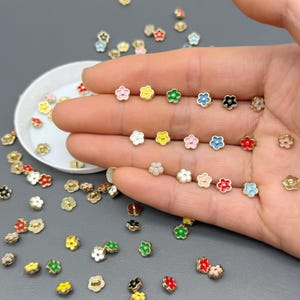 Puede incluir: Una colección de pequeños pendientes de botón con forma de flor en varios colores, incluyendo rosa, amarillo, verde, azul, blanco y rojo. Los pendientes tienen una base dorada y están dispersos sobre una superficie gris.
