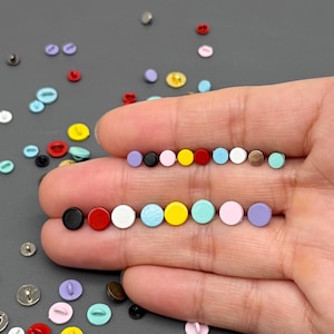 Puede incluir: Una variedad de pequeños botones coloridos en varios tonos de rosa, azul, amarillo, rojo, negro y marrón. Los botones están dispuestos en filas y dispersos sobre una superficie gris. Los botones son pequeños, redondos y tienen un acabado brillante.