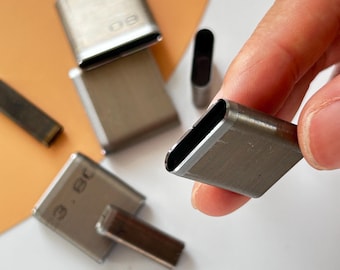 May include: Close-up of several silver metal leather craft tools. One tool is held by a hand. The tools are rectangular with rounded ends and have measurements etched on them. The tools are used for leather working.