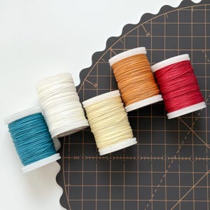 Puede incluir: Vista superior de seis carretes de hilo en varios colores: turquesa, blanco roto, amarillo, naranja y rojo. Los carretes están sobre una esterilla de corte negra con un patrón de cuadrícula. El hilo está enrollado en carretes blancos.