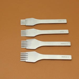 May include: Four silver leathercraft tools with multiple prongs. Each tool has a handle and is labeled with a measurement in millimeters: 2.7MM, 3.0MM, 3.38MM, and 3.85MM. The tools are arranged on a brown surface.