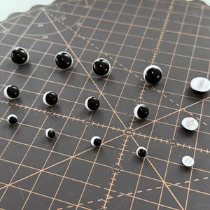 Puede incluir: Una colección de ojos de muñeca en blanco y negro de varios tamaños. Los ojos tienen una pupila negra brillante y un iris blanco en forma de media luna. Están dispuestos sobre una esterilla de corte gris oscuro con líneas de cuadrícula.