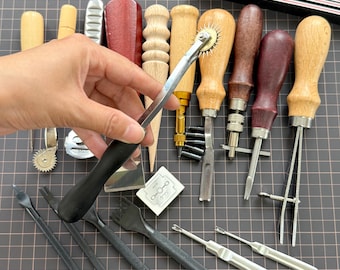 Può includere: Una collezione di strumenti per la lavorazione della pelle, tra cui vari lesine, smussatori per bordi e scanalatori per cuciture. Gli strumenti hanno manici in legno e metallo, con una mano che tiene uno strumento con manico nero al centro. Sono visibili anche altri strumenti.