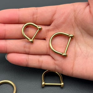 May include: Several brass-colored D-rings are displayed. The D-rings have a rounded top and a straight bar across the bottom. The metal has a shiny finish. The D-rings are likely used for leathercraft, bag making, or other crafting projects.