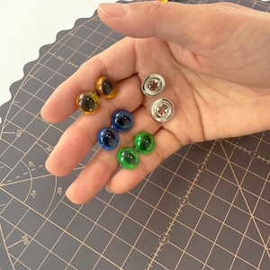 May include: A collection of colorful craft eyes in yellow, blue, and green, along with silver metal fasteners. The eyes have a glossy, rounded appearance, and are held in a hand against a gray cutting mat.