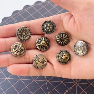 Może przedstawiać: Kolekcja metalowych guzików w stylu antycznym w odcieniach brązu i srebra. Guziki mają różne wzory, w tym motywy kwiatowe, fleur-de-lis i głowę lwa. Jeden guzik ma napis "Tschou".