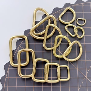 May include: A collection of various sizes of gold-colored metal D-rings and rectangular rings. The rings are arranged on a black cutting mat with a grid pattern. The D-rings are in different sizes, and some are interlocked.