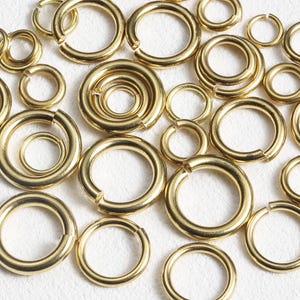 May include: A collection of gold-colored jump rings of various sizes, scattered on a white surface. The rings are open, ready for use in jewelry making or other crafts. The lighting highlights the metallic sheen of the rings.