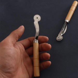 Könnte beinhalten: Zwei Schneiderädchen mit Holzgriffen und Metallrädern. Eines wird in einer Hand gehalten, das andere liegt auf einer dunklen Oberfläche. Die Räder haben eine Reihe kleiner, spitzer Zähne am Rand.