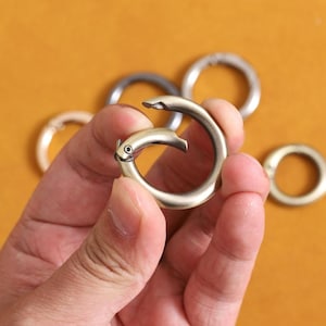 May include: A hand holding a bronze-colored, bird-shaped carabiner clip. The clip is open, revealing a circular design. Other metal rings are visible in the background against a yellow-orange backdrop. The clip has a small, round eye detail.