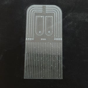 Peut inclure: Un gabarit en acrylique transparent dot&eacute; d'une grille de petits trous et de pistes incurv&eacute;es. L'outil porte les inscriptions 3mm, 10, 12, 15, 20, 25, 30, 35, 40 et 45. Il pr&eacute;sente une base rectangulaire avec un bord sup&eacute;rieur arrondi.