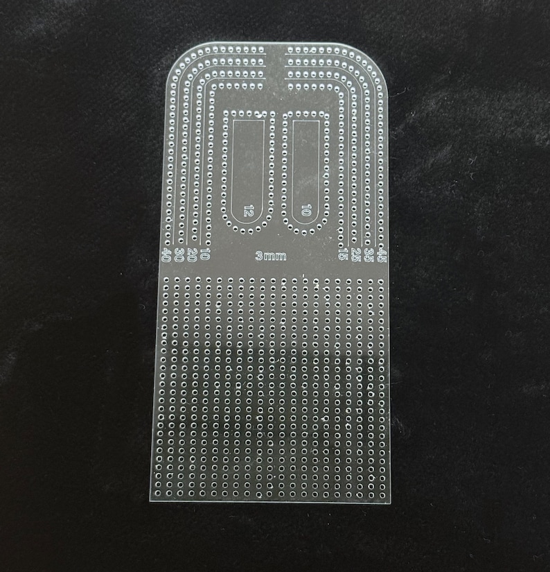 Peut inclure: Un gabarit en acrylique transparent dot&eacute; d'une grille de petits trous et de pistes incurv&eacute;es. L'outil porte les inscriptions 3mm, 10, 12, 15, 20, 25, 30, 35, 40 et 45. Il pr&eacute;sente une base rectangulaire avec un bord sup&eacute;rieur arrondi.