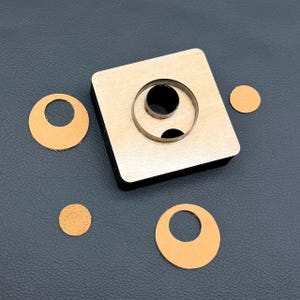 May include: A square wooden leather cutting die with a circular metal blade, accompanied by two tan leather circles with centre cutouts and two small solid tan leather circles, all arranged on a dark grey surface.