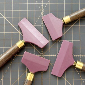 K&ouml;nnte beinhalten: Vier mauvefarbene Rakel mit dunklen Holzgriffen und Messingzwingen sind auf einer schwarzen Rasteroberfl&auml;che angeordnet. Die Rakel haben ein flaches, abgewinkeltes Klingendesign, ideal f&uuml;r verschiedene Bastel- oder Reinigungsaufgaben. Die Griffe sind dunkelbraun.
