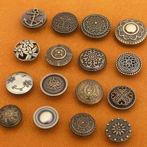 Op de afbeelding: Verzameling antieke bronzen knopen met diverse ontwerpen, waaronder een anker, bloempatronen en ingewikkelde geometrische vormen. De knopen zijn rond en hebben een vintage uitstraling, perfect voor naai- of knutselprojecten.