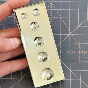 May include: A rectangular, polished silver metal object with five circular, raised, button-like embellishments. The buttons vary in size, with the largest at the bottom. The object is held against a dark gray grid background.