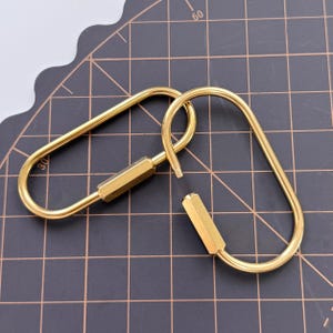 Puede incluir: Dos mosquetones dorados de forma ovalada con un mecanismo de cierre de rosca. Los mosquetones son de metal y tienen un acabado brillante. El fondo es una superficie oscura con un patrón de cuadrícula.