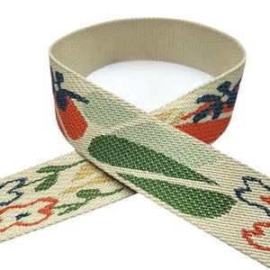 May include: A beige woven elastic ribbon featuring a repeating pattern of stylized flowers and leaves in shades of dark blue, muted green, and orange. The fabric has a textured, durable appearance with a consistent weave throughout the material.